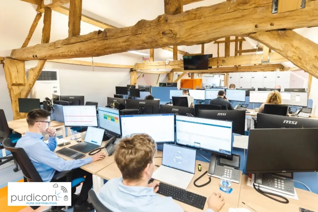 Dipendenti al lavoro alle loro scrivanie, circondati da più monitor, computer portatili e telefoni fissi, in un ufficio open space con travi a vista.